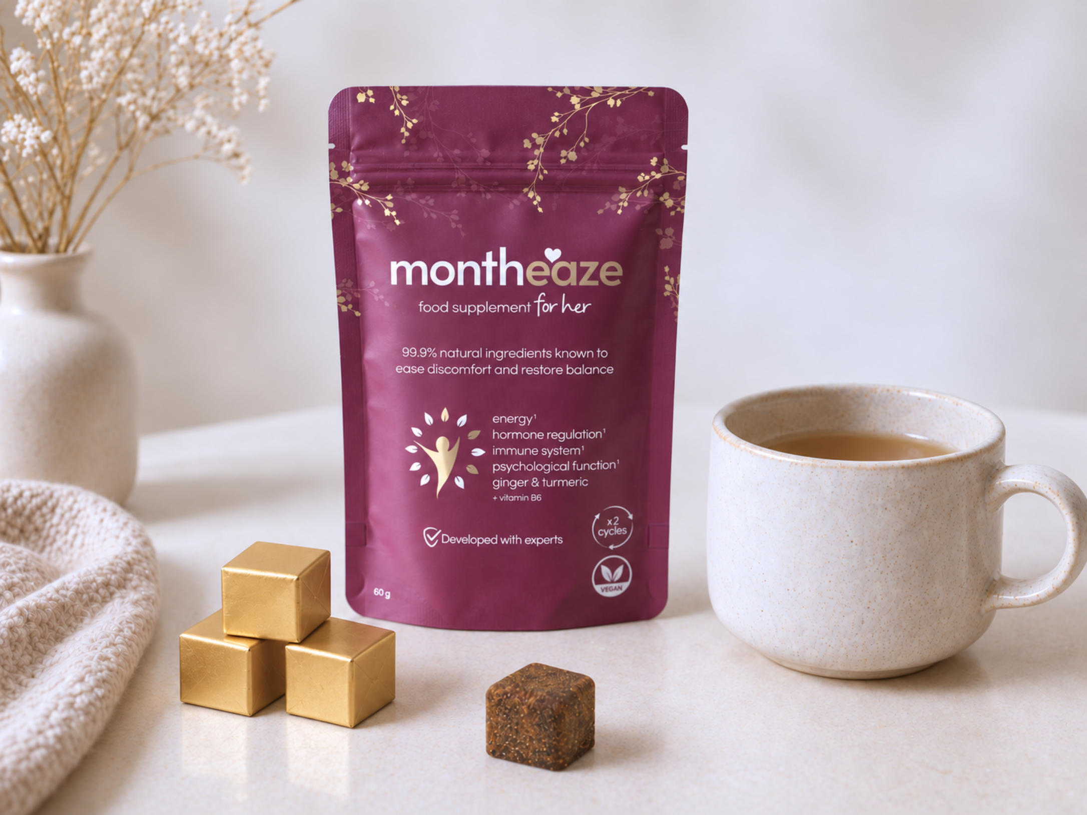 A stand-up pouch of monthaze food supplement for her in a deep plum purple color with gold floral accents, displayed on a light marble surface next to a cup of tea, gold-wrapped chocolates, and a textured cream blanket. Purple package of 'montheaze' on a white surface with a cup and cubes.