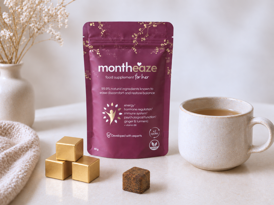 A stand-up pouch of monthaze food supplement for her in a deep plum purple color with gold floral accents, displayed on a light marble surface next to a cup of tea, gold-wrapped chocolates, and a textured cream blanket. Purple package of 'montheaze' on a white surface with a cup and cubes.
